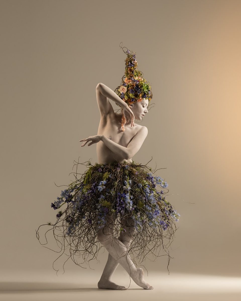 A dancer on pointe in a skirt of lavender and greenery, with a tall floral headpiece.