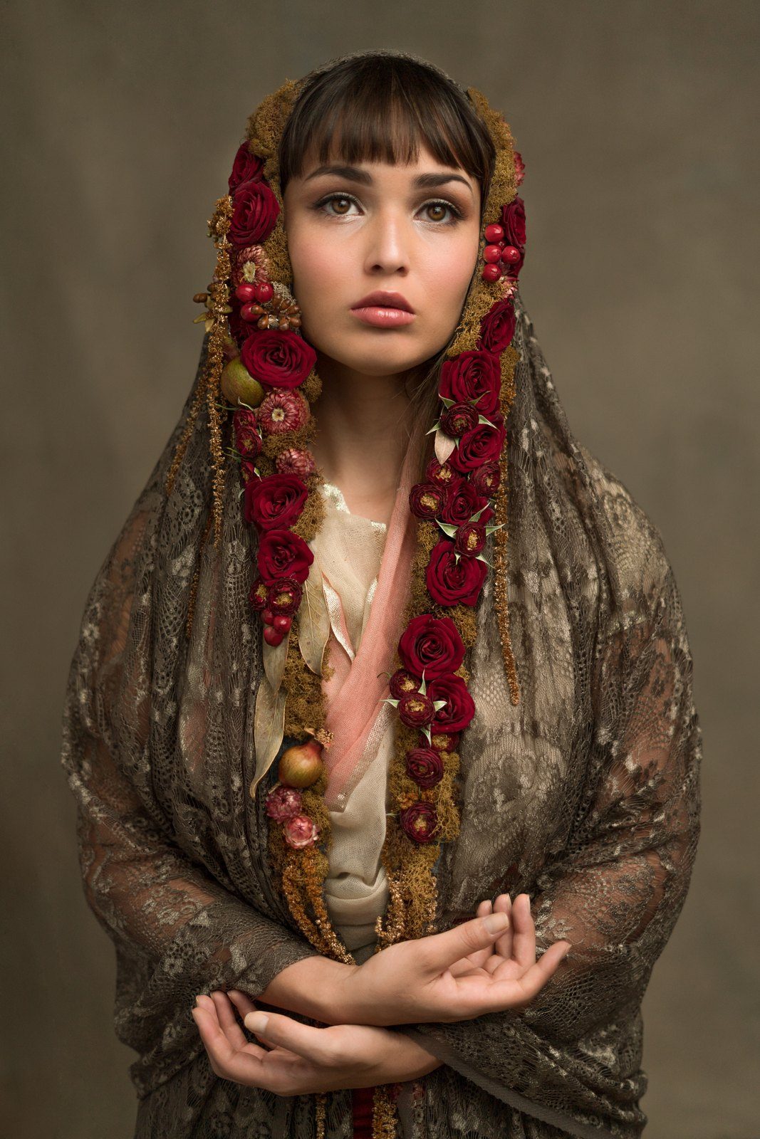 A gold lace shawl veiled over the head, edged with a garland of red roses. Hands are folded.